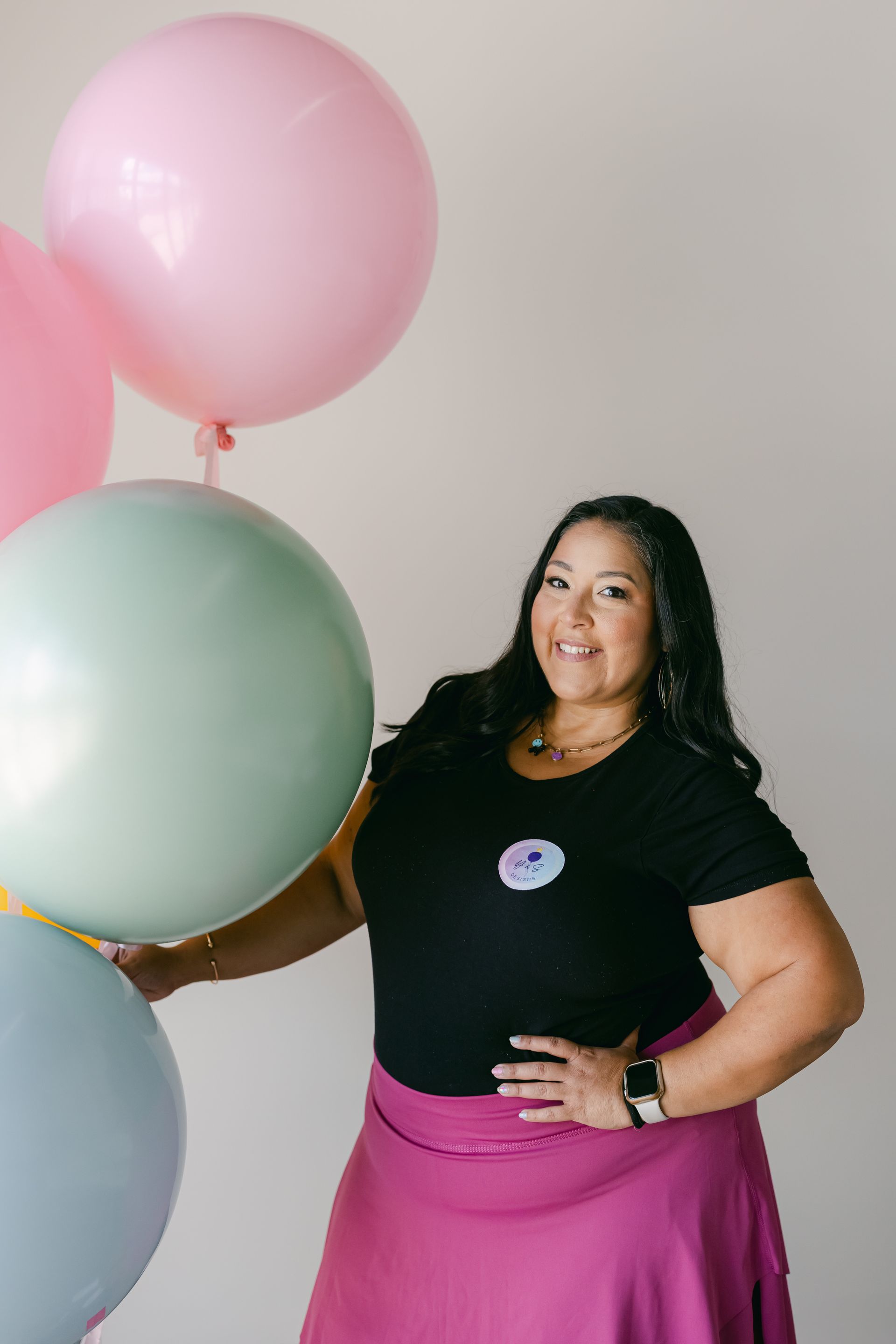 Woman in black top and pink skirt standing with pastel balloons, smiling at the camera.