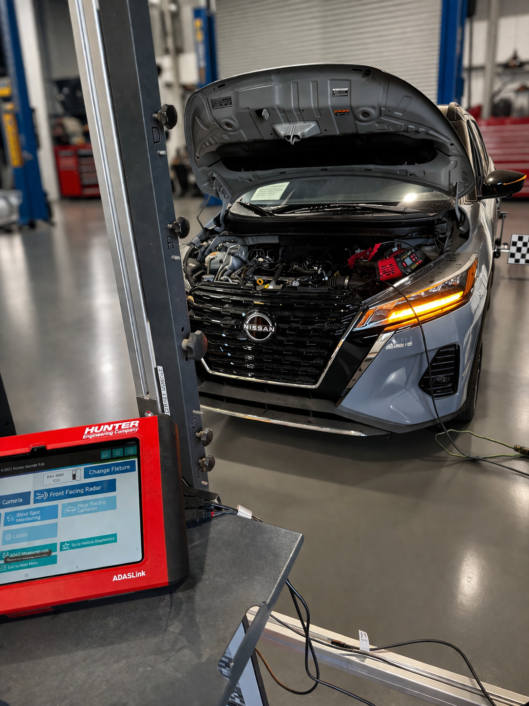 Black car with hood open in a workshop, seen near a tablet on a stand.