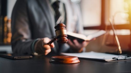 Judge’s gavel on a desk with a person in a suit holding a pen beside documents and a laptop
