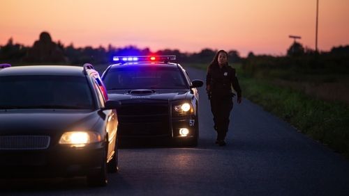 Police cars with flashing lights stop on a road at dusk, with an officer walking beside them.