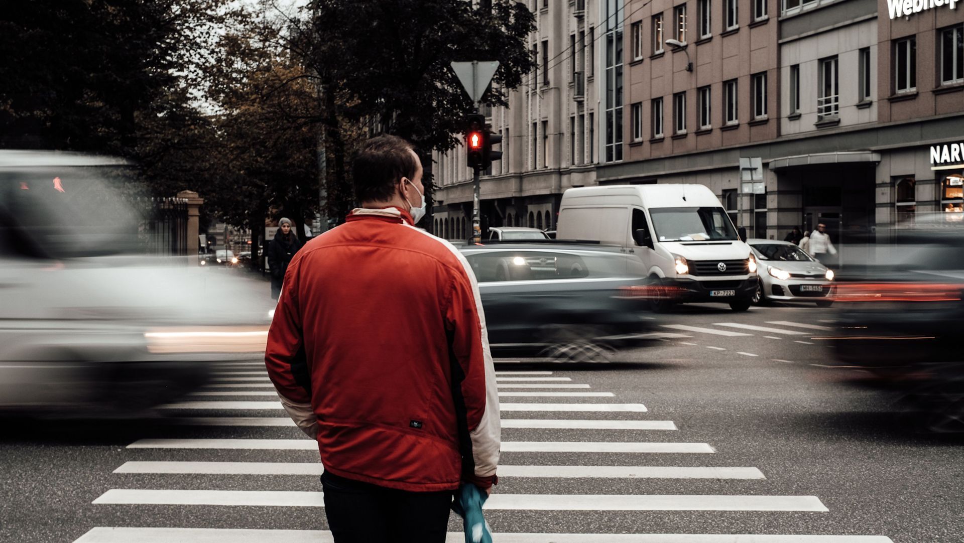 Person in a red jacket standing at a crosswalk with blurred traffic passing by on a city street