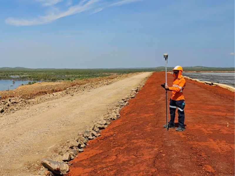 Survey worker in orange safety gear using a GPS pole on a dirt road beside red earth and water.