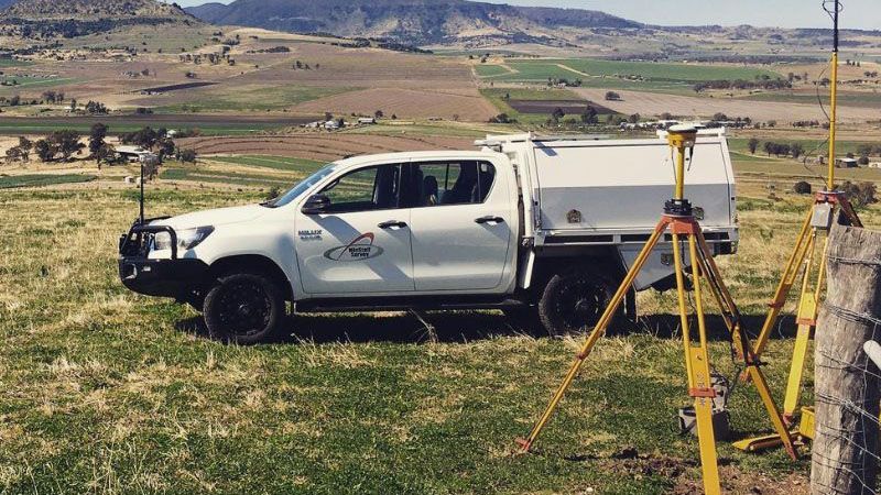 White utility truck parked beside survey equipment in a grassy field with mountains in the background