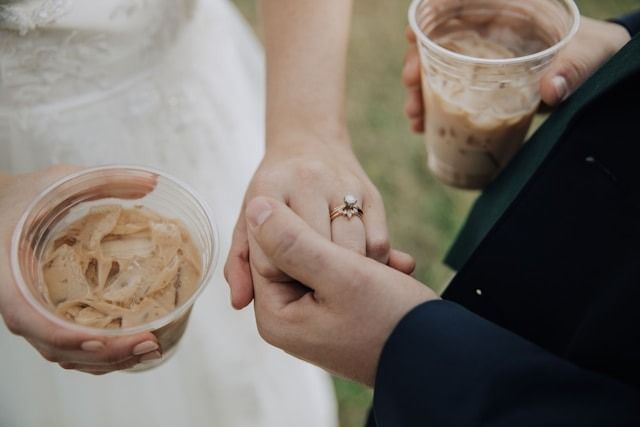 Two people holding hands with plastic cups of iced coffee, one in a white dress and one in a dark suit