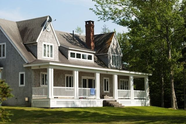 Gray house with white porch and gabled roof, surrounded by green trees and lawn
