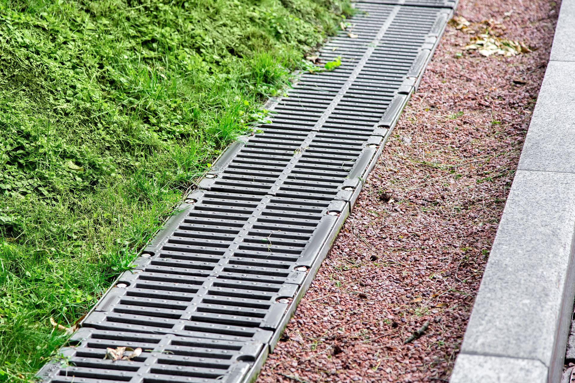 Metal drainage grate beside a grassy slope and gravel path along a concrete curb