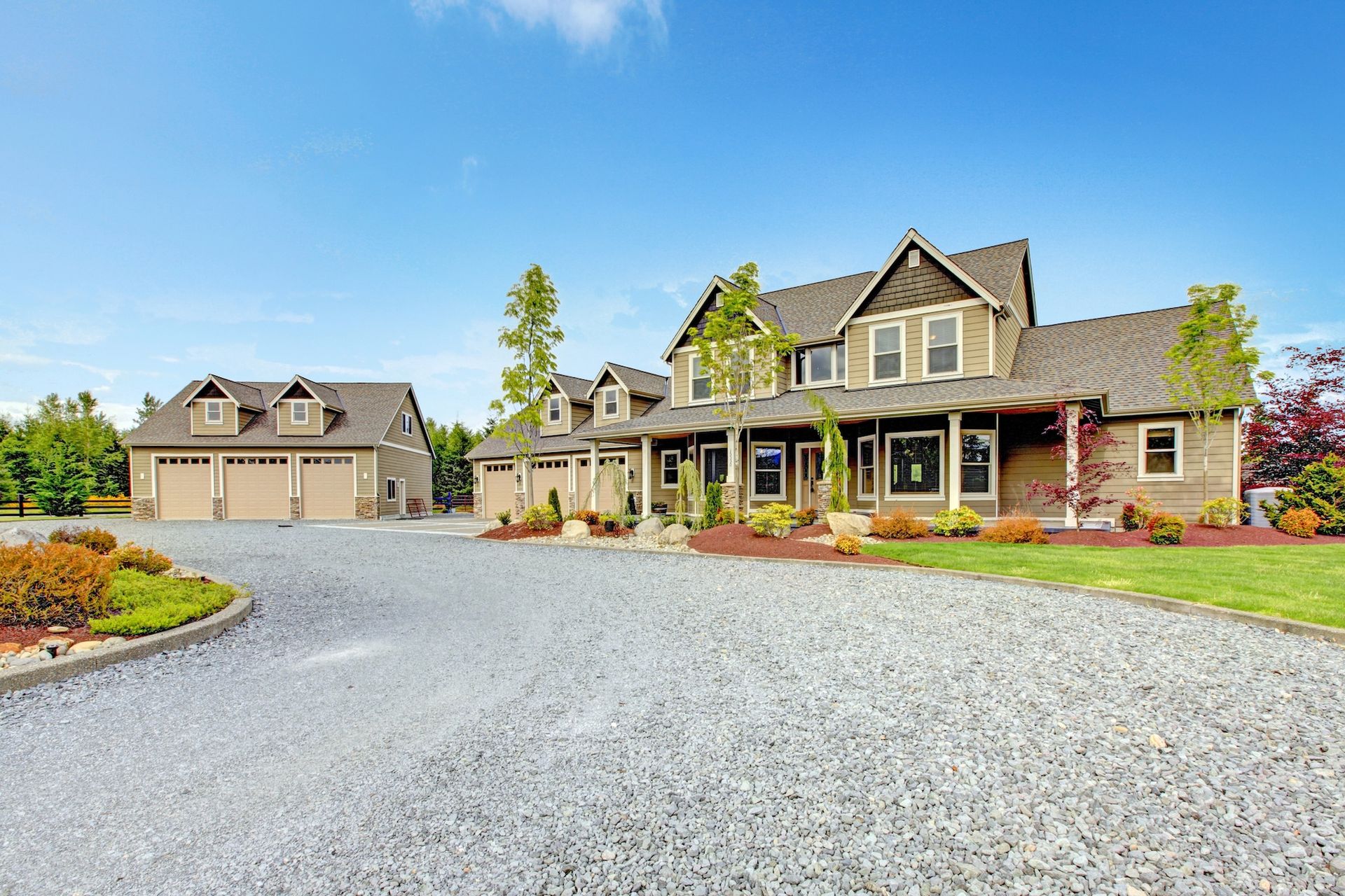 Large gray suburban house with a gravel driveway, front porch, and detached garage under a blue sky