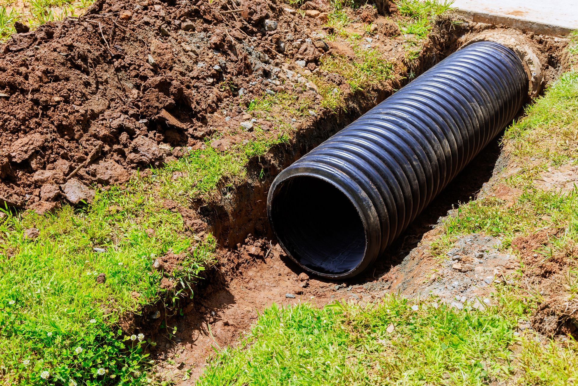 Black corrugated drainage pipe in a dirt trench beside green grass, with soil piled nearby