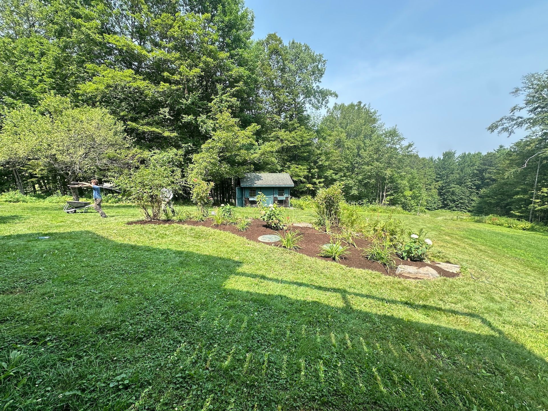 Sunny garden with lush green lawn, flower beds, mature trees, and a small gazebo in the background