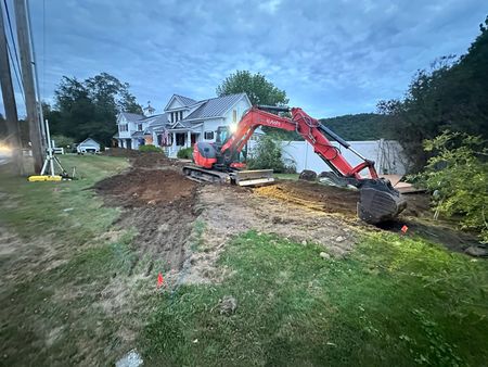 Excavator digging a trench beside a house at dusk, with dirt piled on the lawn.