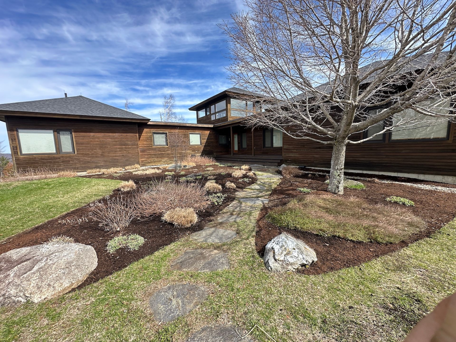 Brick house with a landscaped front yard, stone walkway, rocks, and a leafless tree under a blue sky