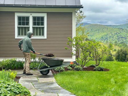 Person pushing a wheelbarrow beside a house, with a green lawn and mountain view.