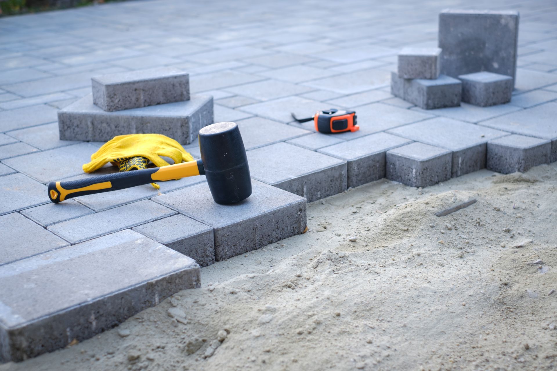 Construction tools on gray paving stones beside a sandy area and stacked concrete blocks