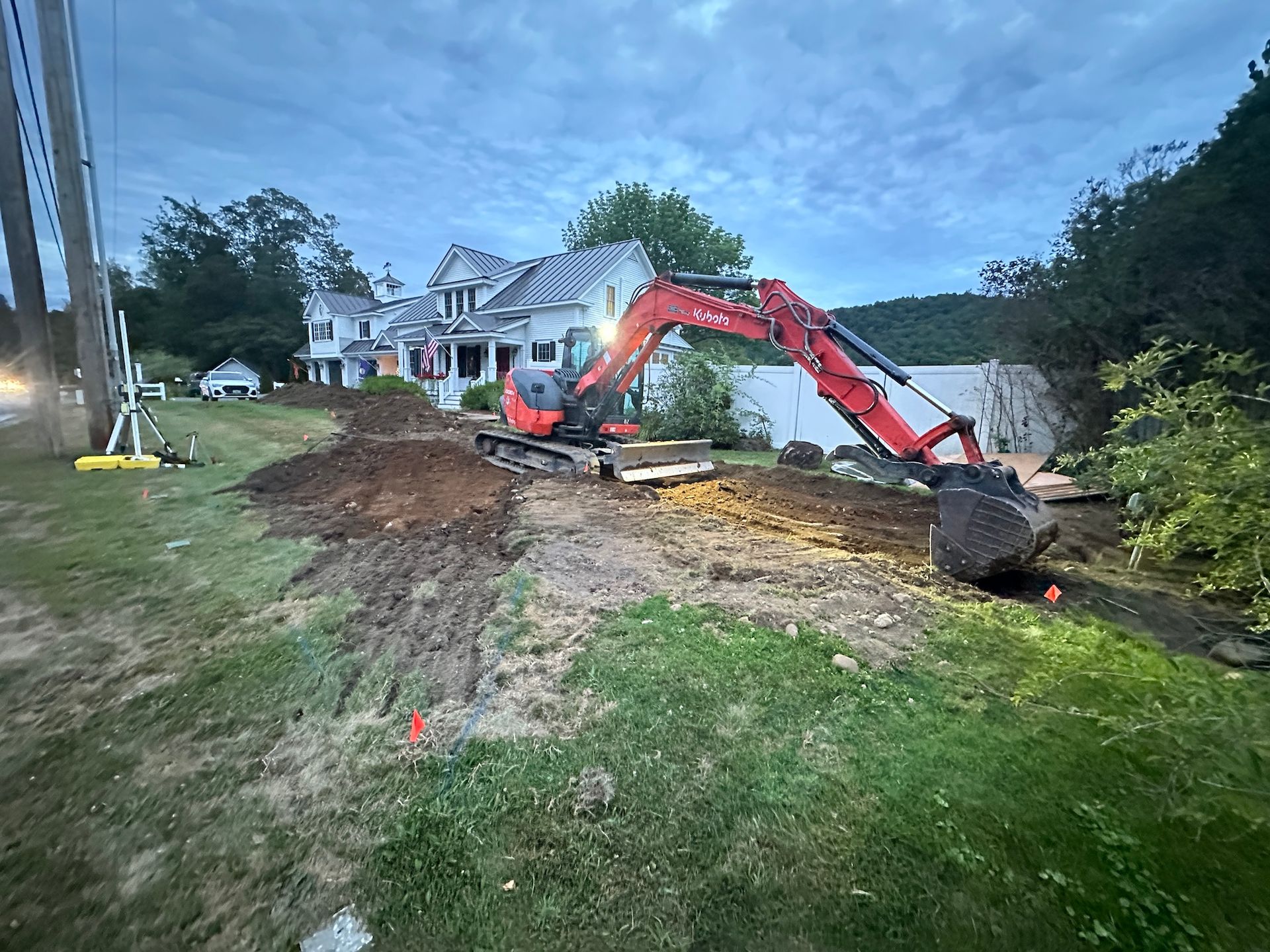 Excavator digging a trench beside a house at dusk, with dirt piled along a grassy yard.