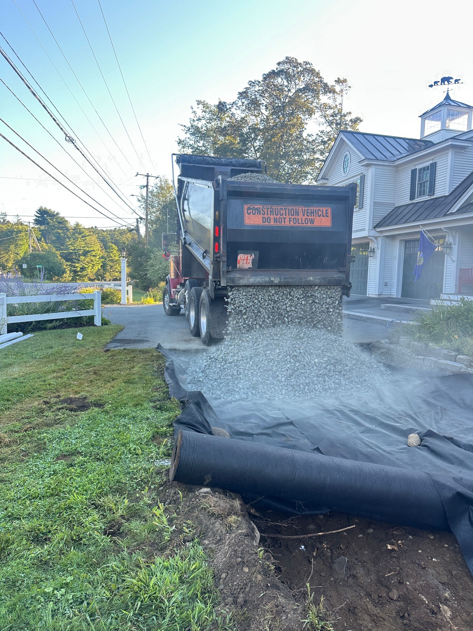 Dump truck pouring gravel into a trench beside a house and grassy roadside.