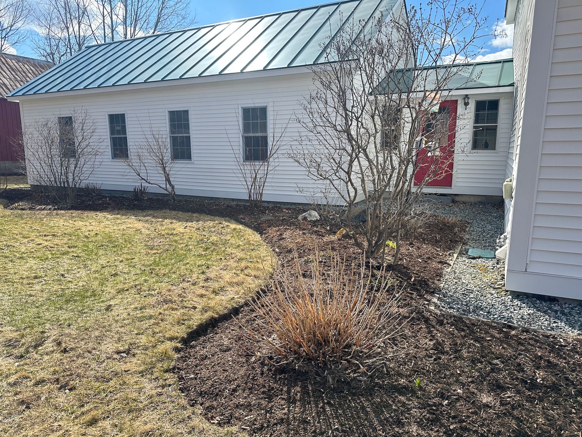 Single-story white house with a teal metal roof, red front door, and a curved mulched garden bed in the yard
