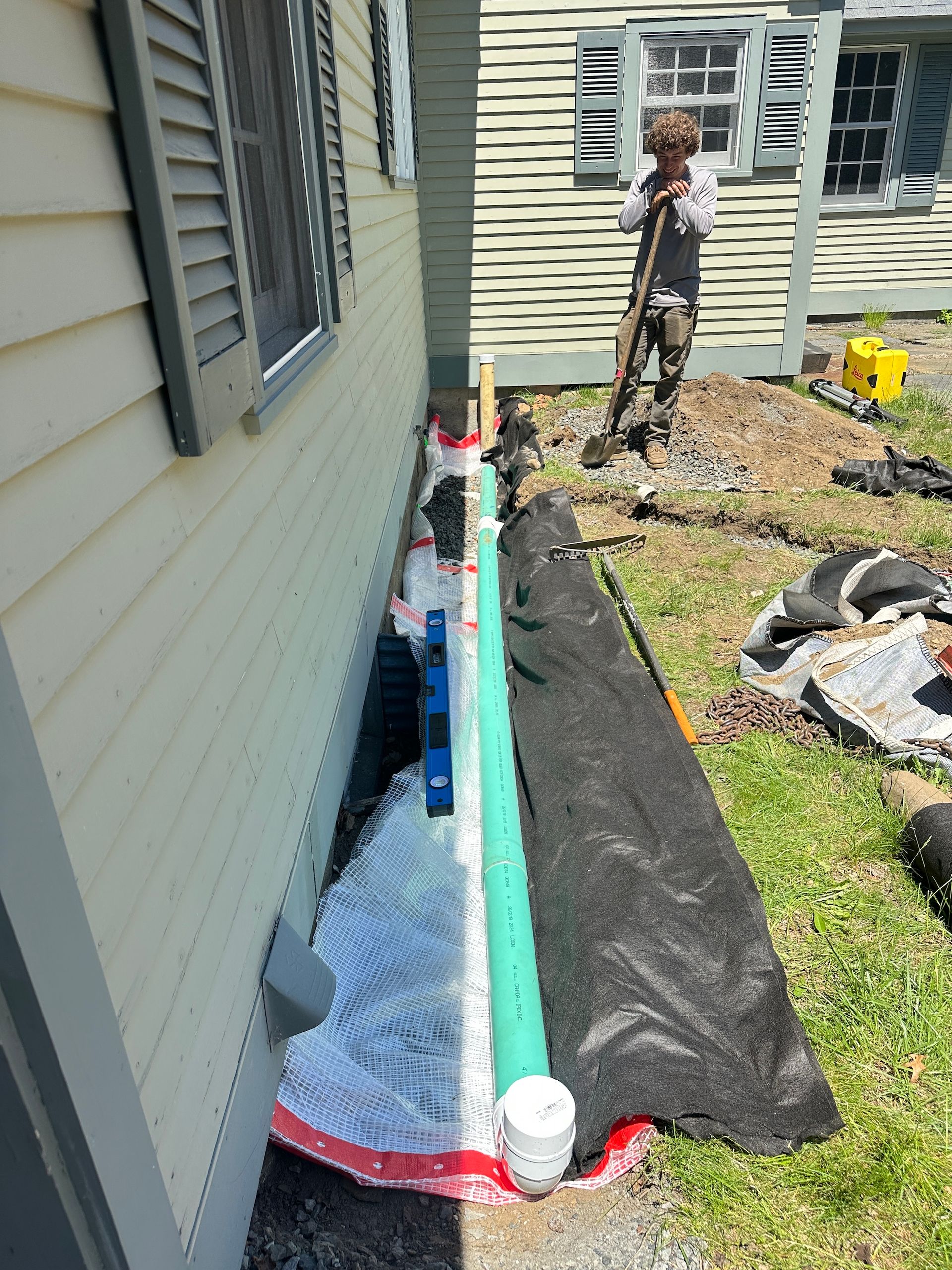 Outdoor utility trench along house wall with insulation wrap, pipes, and a worker standing nearby