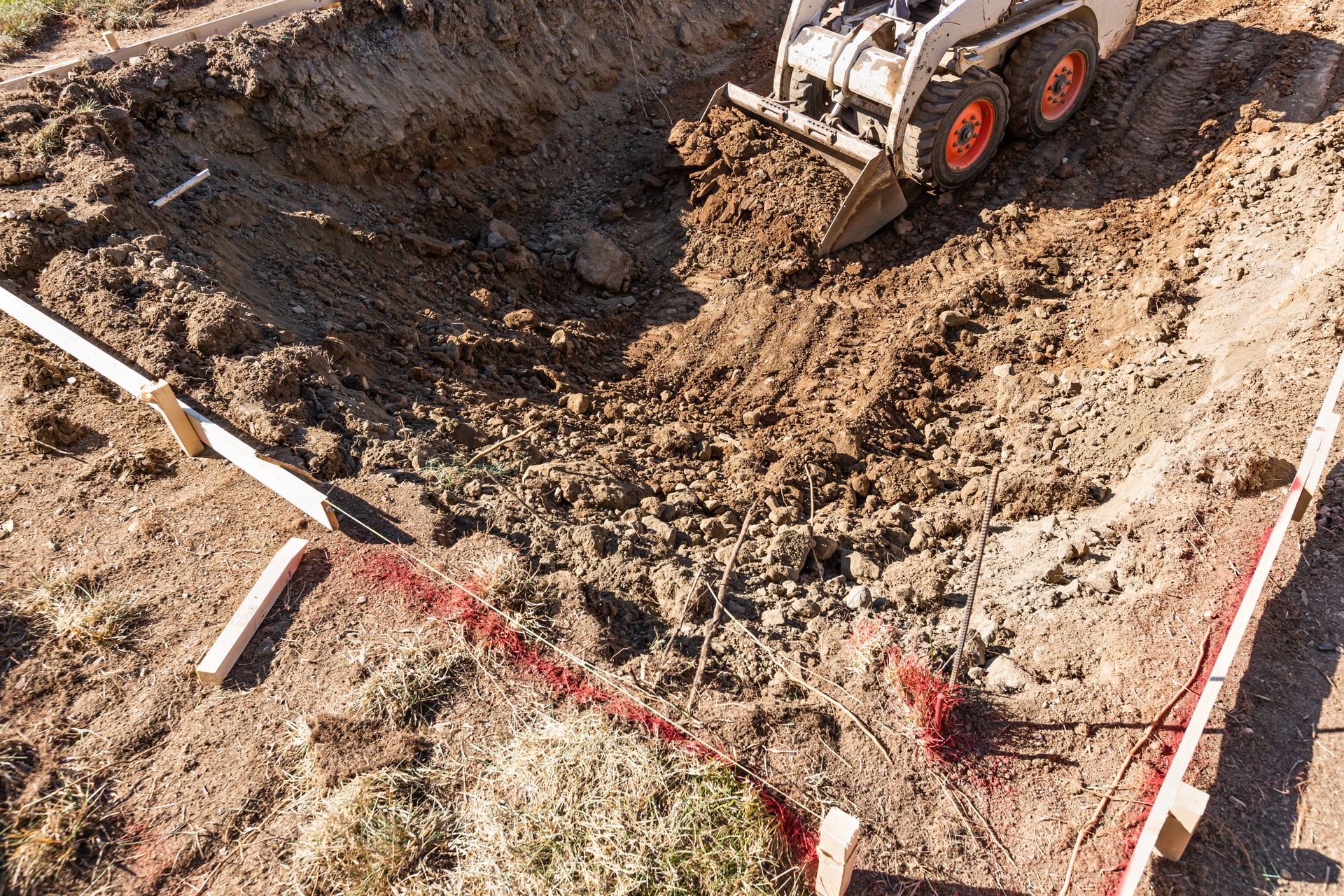 Excavator digging a muddy trench at a construction site, marked with red and white caution tape.