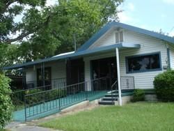 Small white house with blue roof, front porch, and wheelchair ramp surrounded by green lawn and trees
