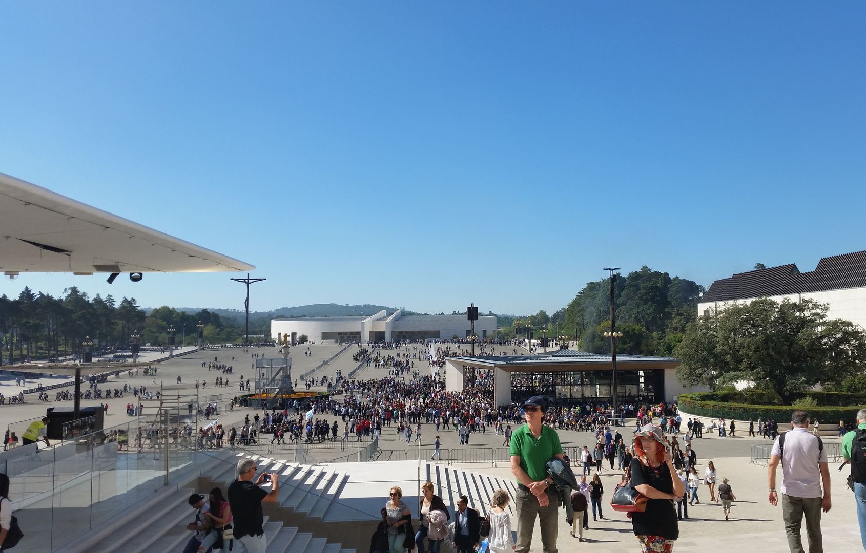 Crowded outdoor plaza by the water under a bright blue sky, with people walking and sitting on steps