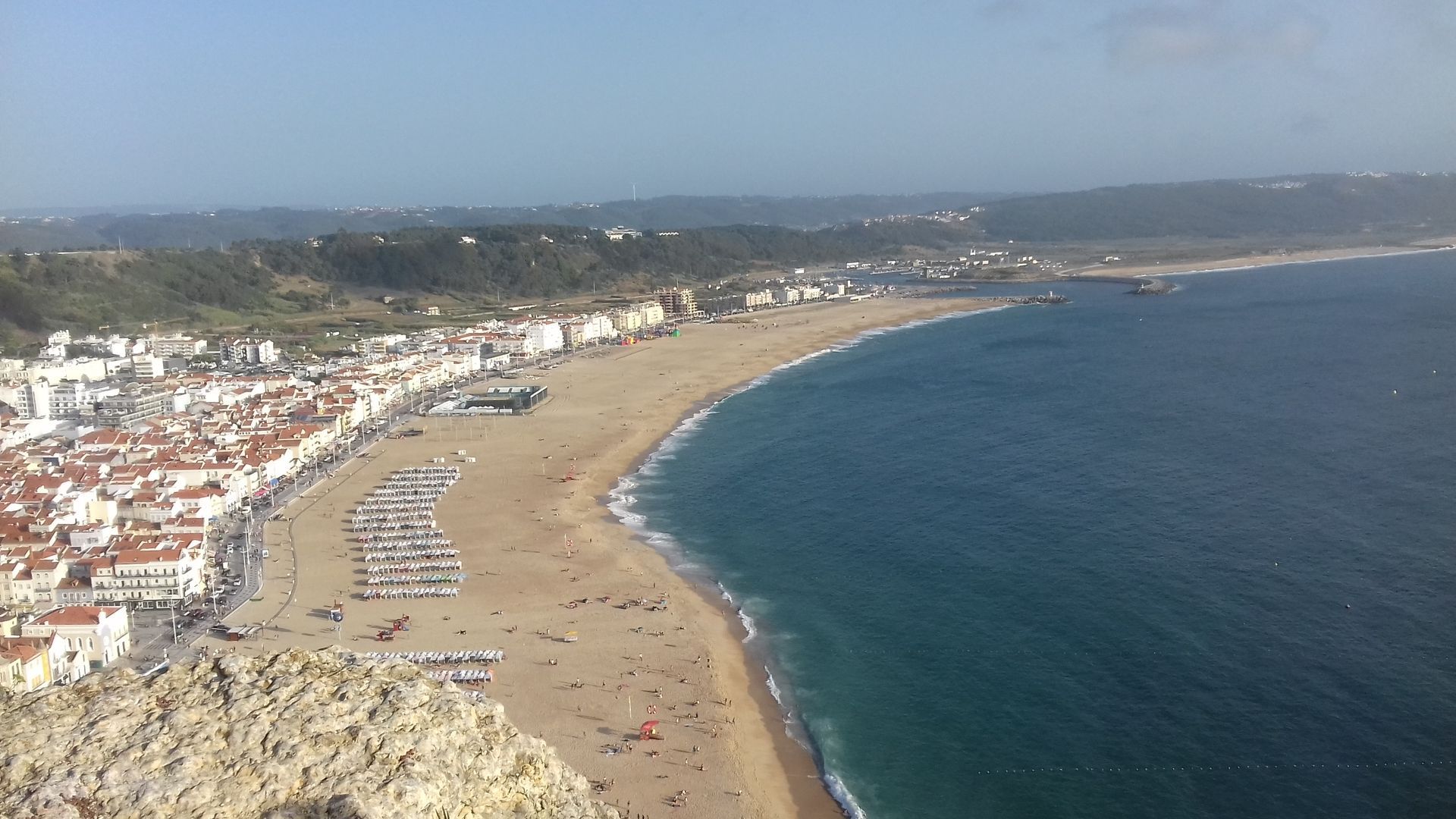 Aerial view of a curved sandy beach beside blue ocean and a coastal town under a hazy sky