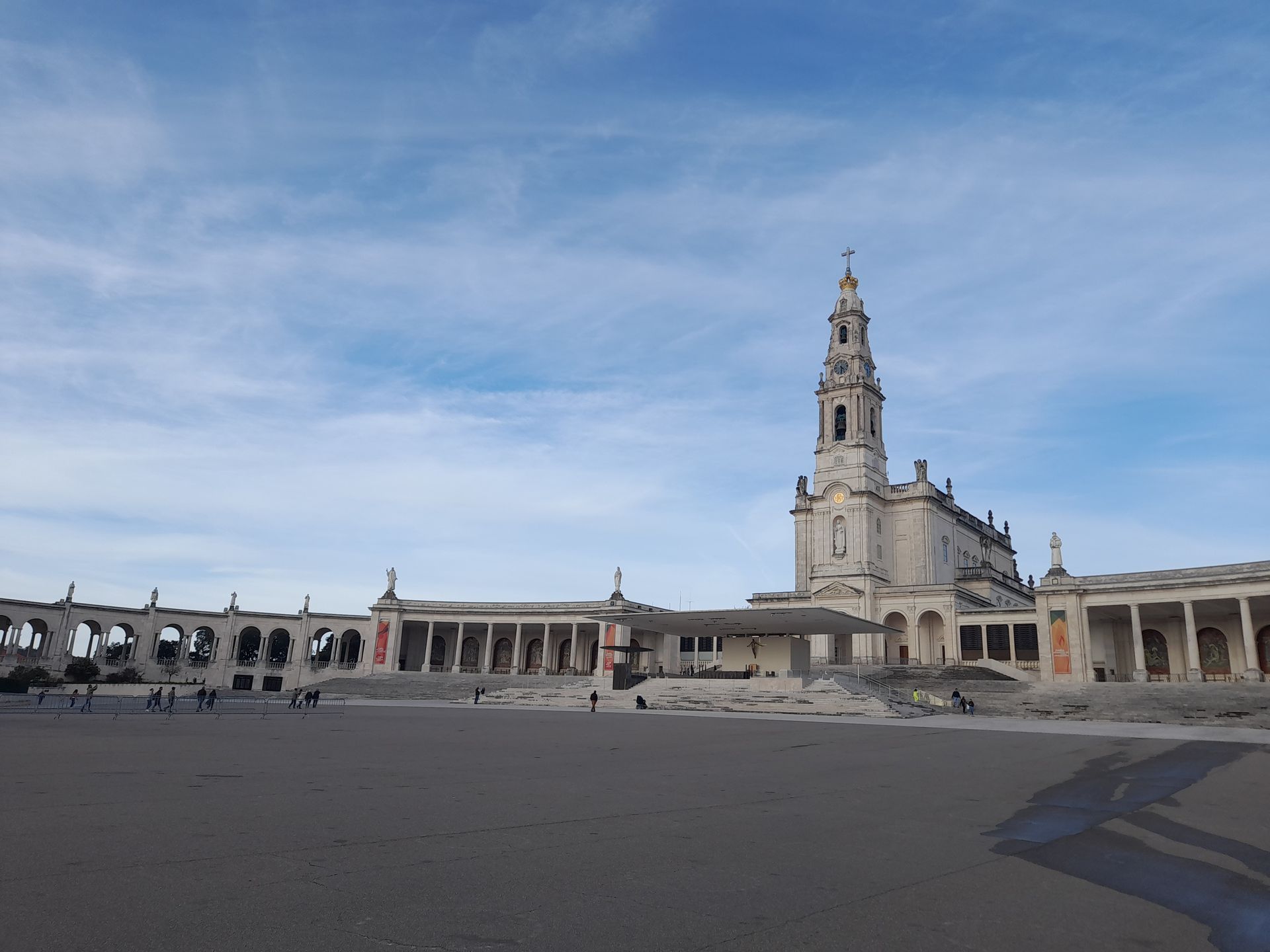 Wide plaza with a historic church tower and colonnaded buildings under a blue sky