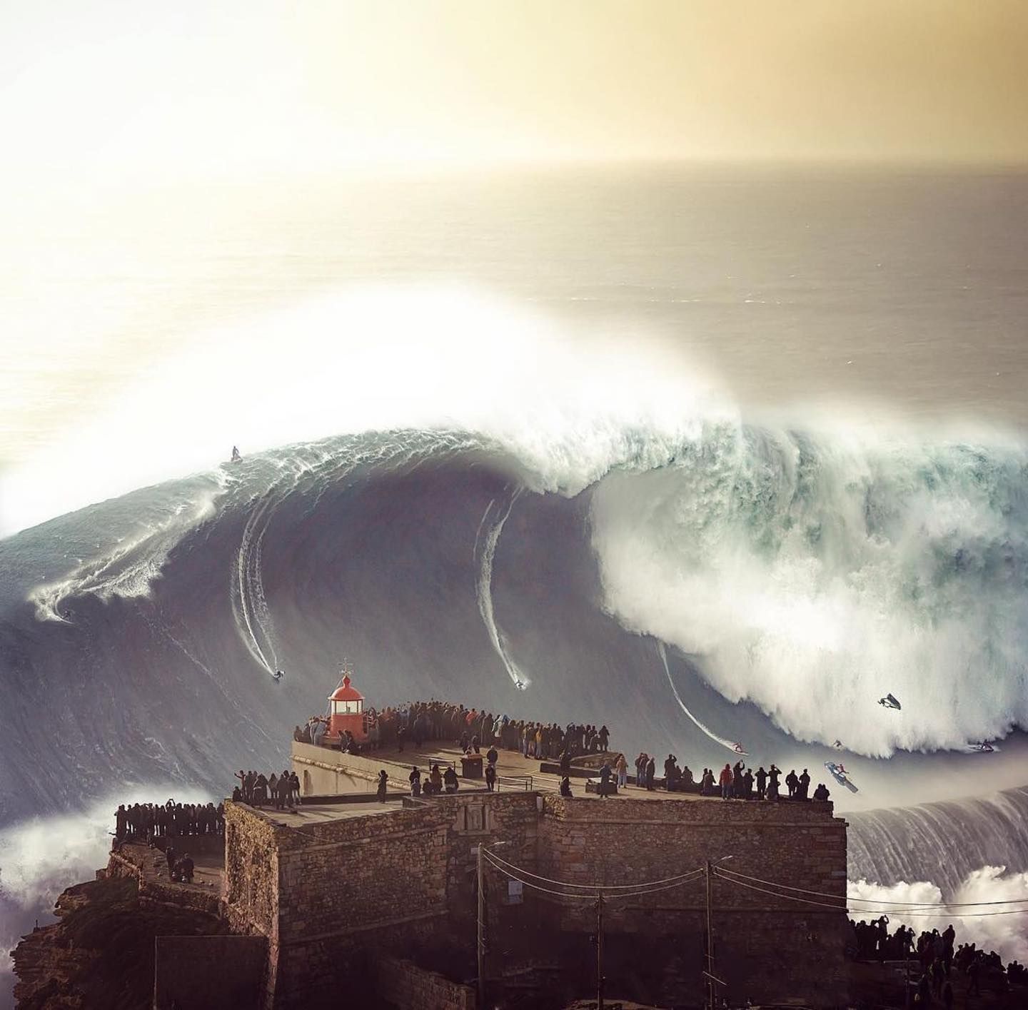 Huge ocean wave crashing behind a crowded fortress under a hazy sky