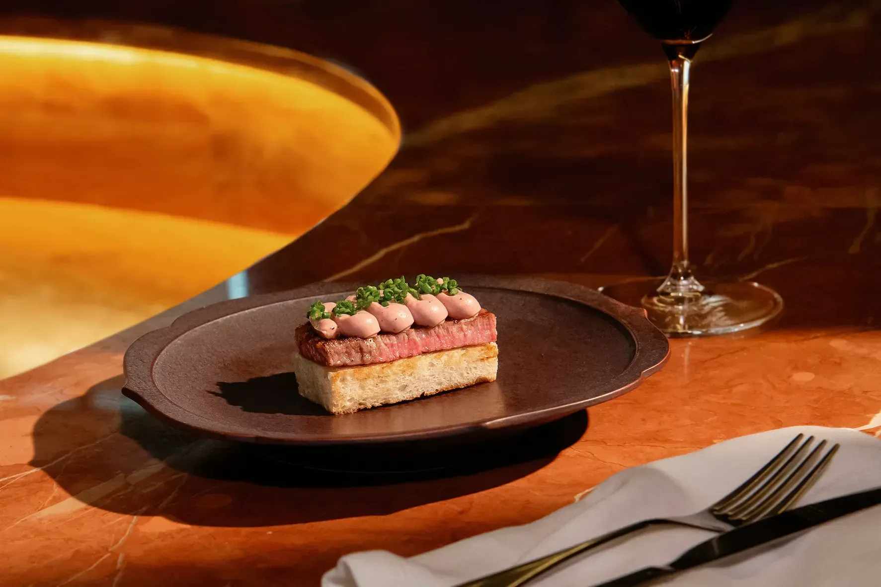A small piece of food is on a plate next to a glass of wine.