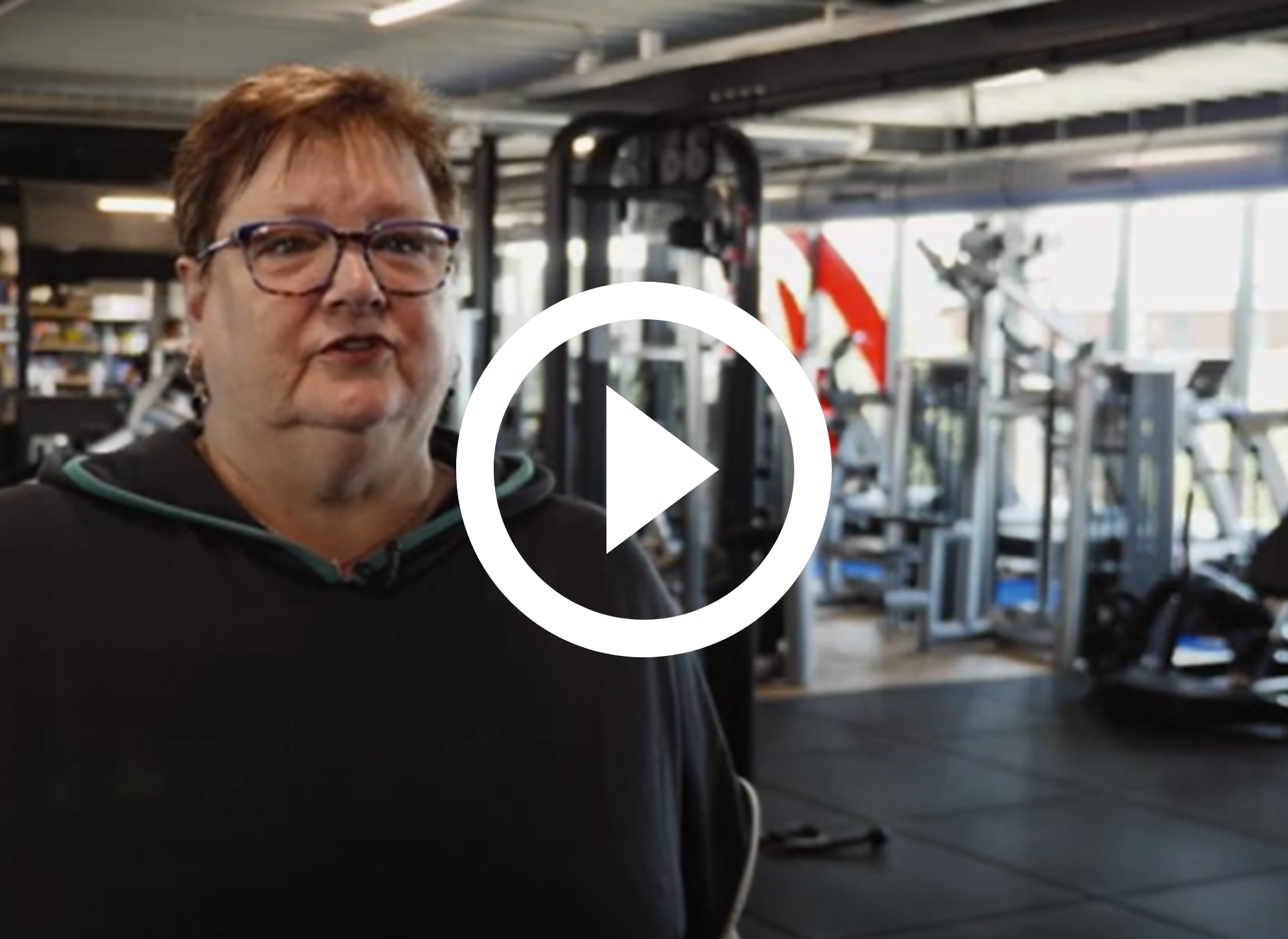 Vrouw staat in een sportschool met fitnessapparatuur en een grote afspeelknop.