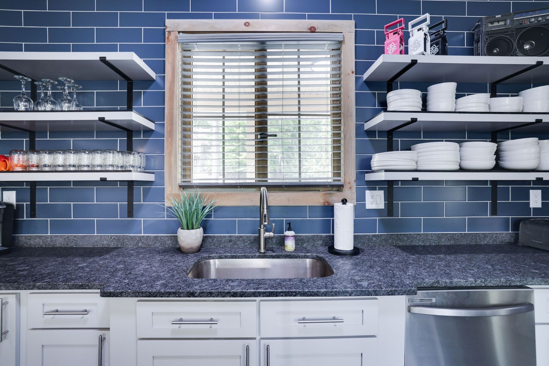 A kitchen with a sink , dishwasher , shelves and a window.