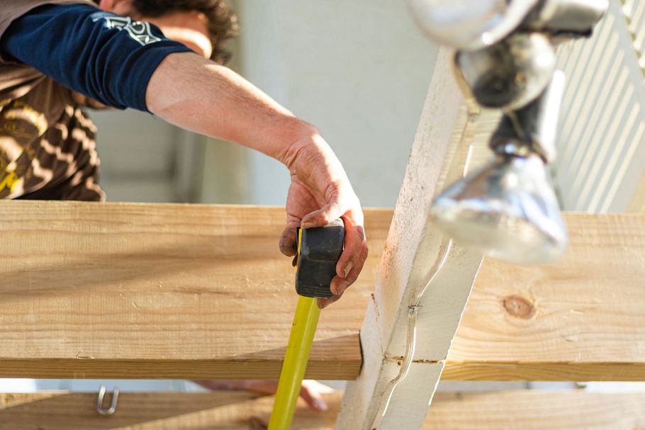 Person measuring a wooden plank on a sawhorse with a yellow tape measure