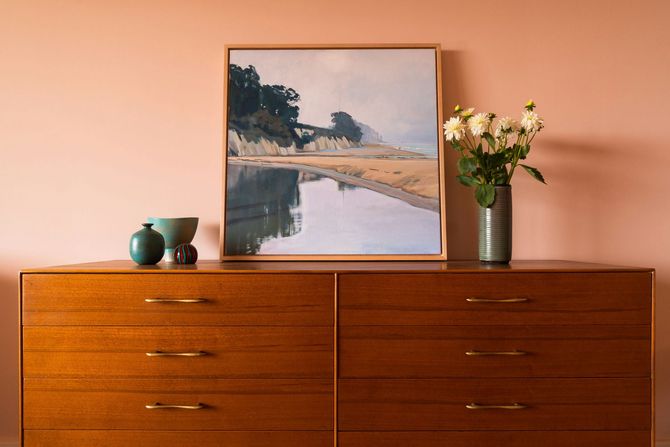 Wooden dresser with framed landscape, vase of flowers, and small green vases against a peach wall.