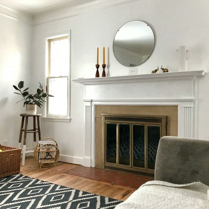 Bright living room with a white fireplace, round mirror, window, plants, and patterned rug.