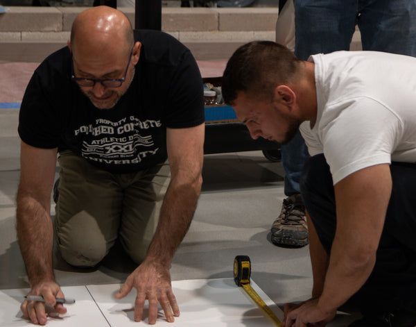 Two men kneel on the floor, measuring and marking a surface with a tape measure and pencil.