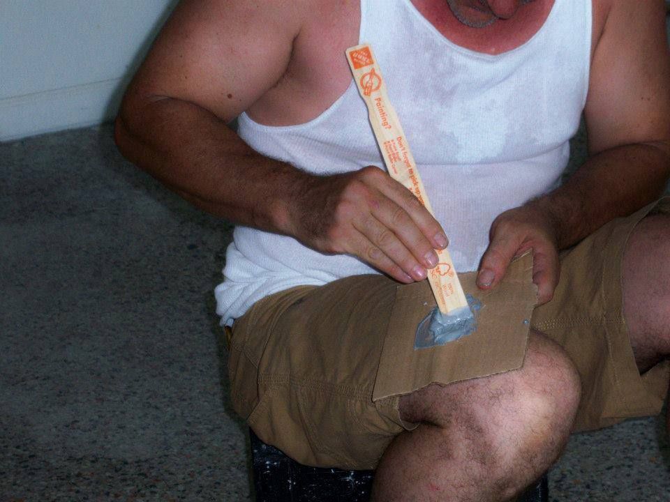 Seated man in a white tank top and tan shorts holding a long box cutter on his lap