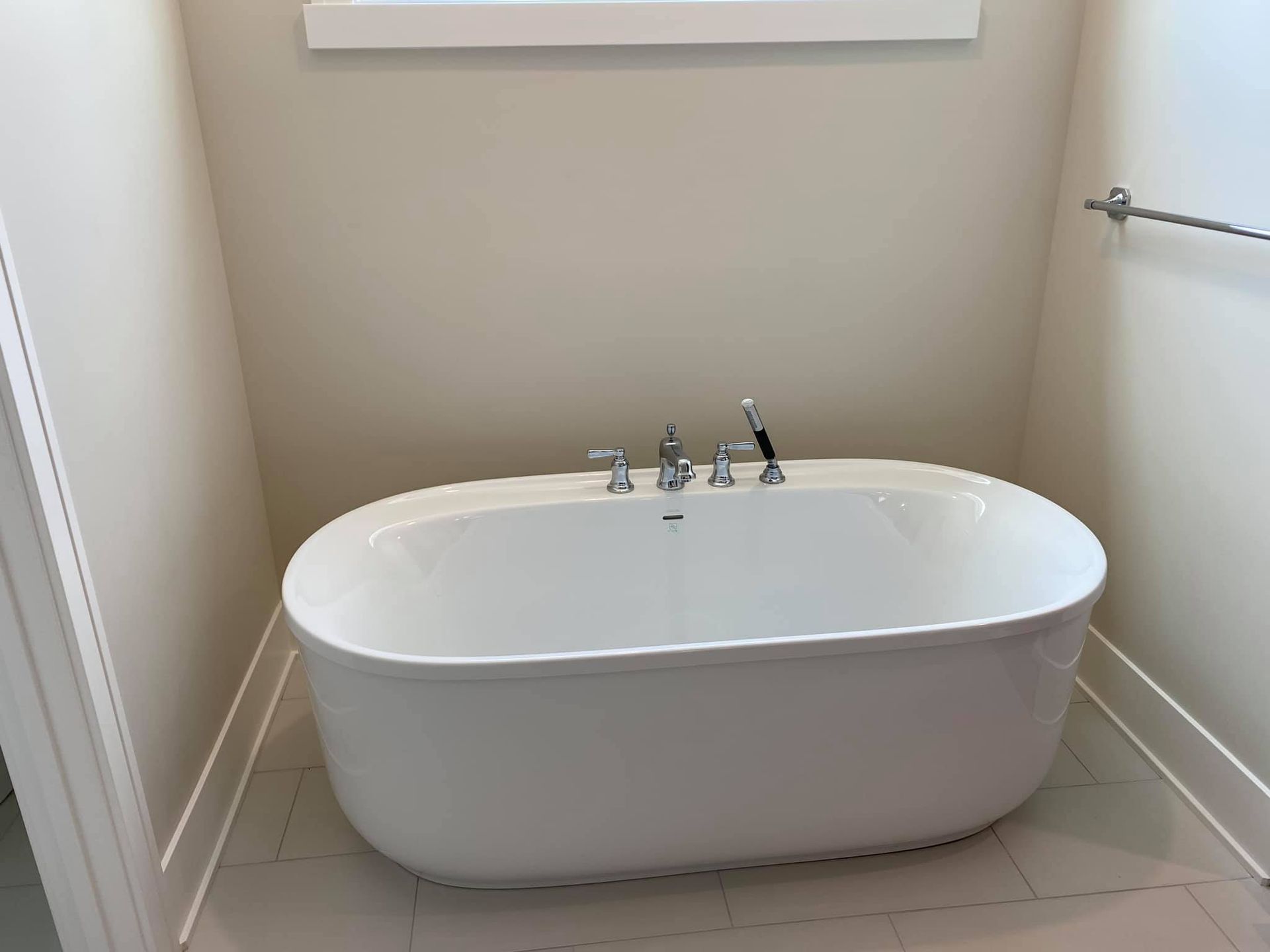 White freestanding bathtub in a small beige bathroom with tiled floor and chrome fixtures