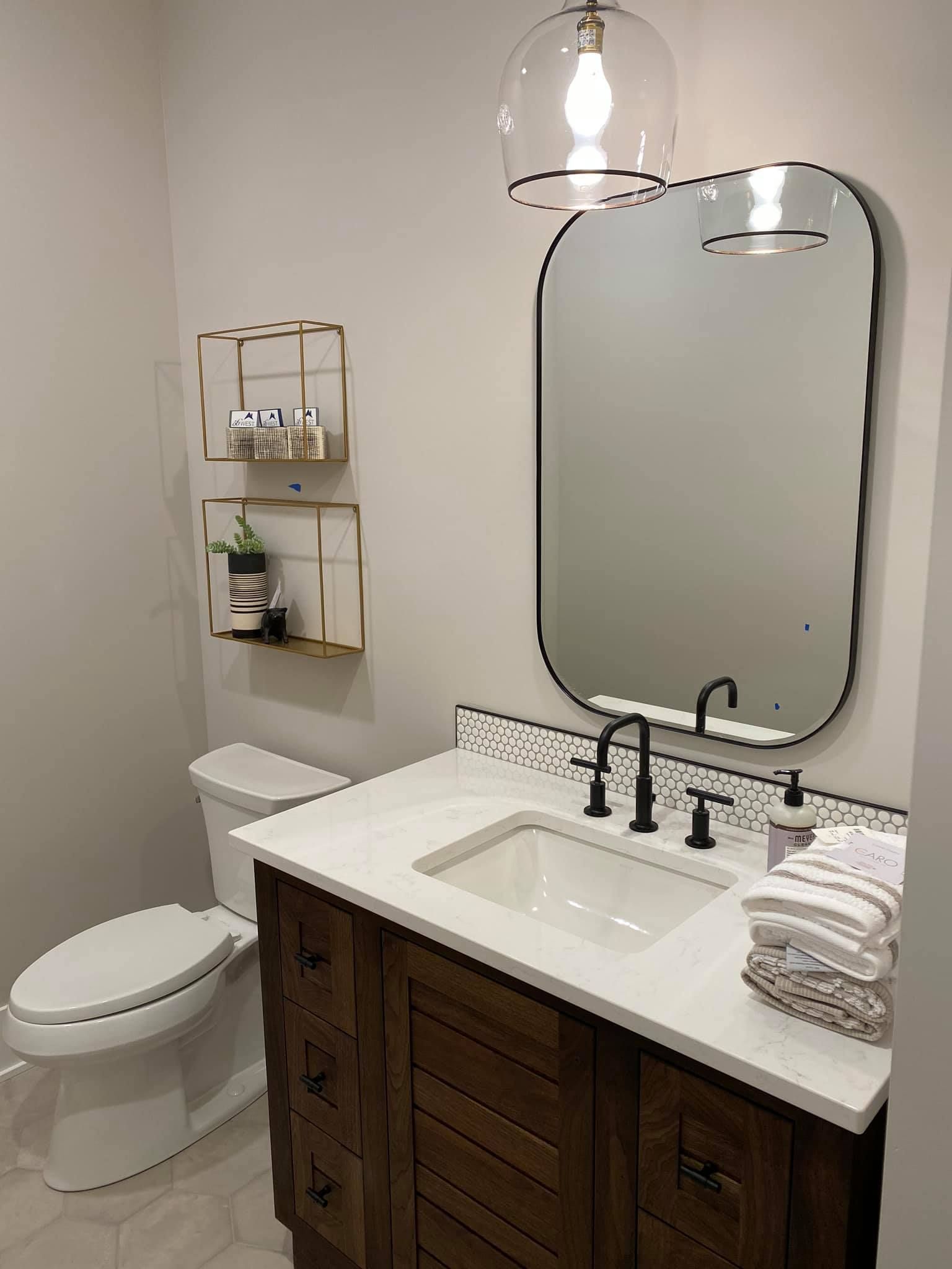 Modern bathroom with white vanity, black fixtures, round mirror, toilet, and wall shelf.