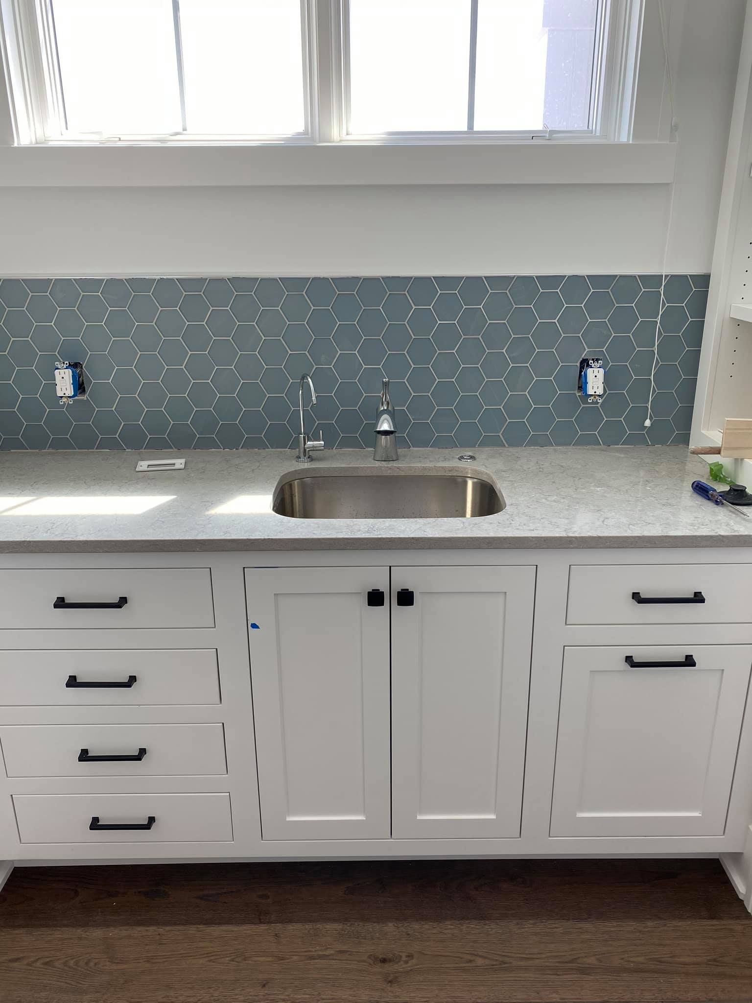 White kitchen with gray mosaic backsplash, stainless sink, and cabinets under a window