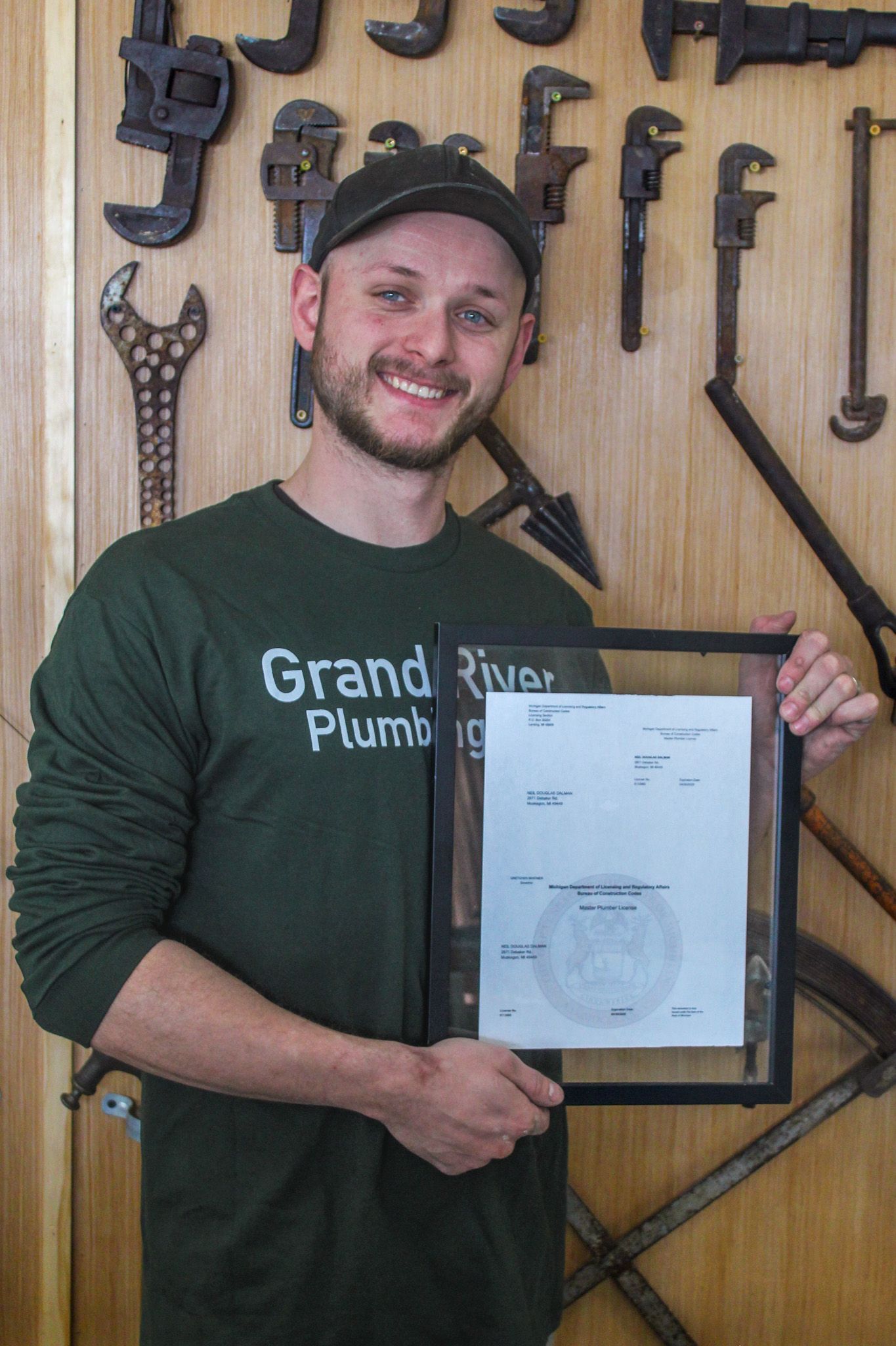 Man in dark green shirt holding a framed certificate in front of a wall of tools and wrenches