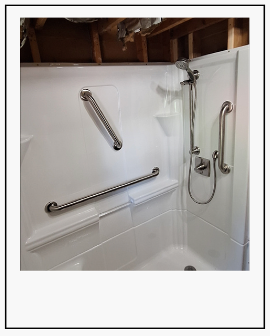 Accessible shower with grab bars and a handheld showerhead in a white bathroom stalled with exposed ceiling framing