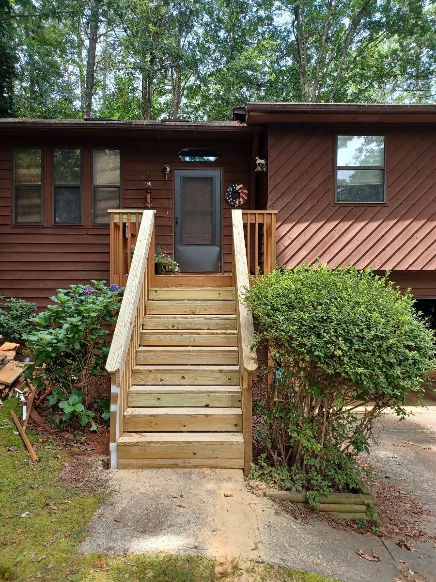 Wooden stairs lead up to a brown house with a screen door. Bushes flank the stairs.
