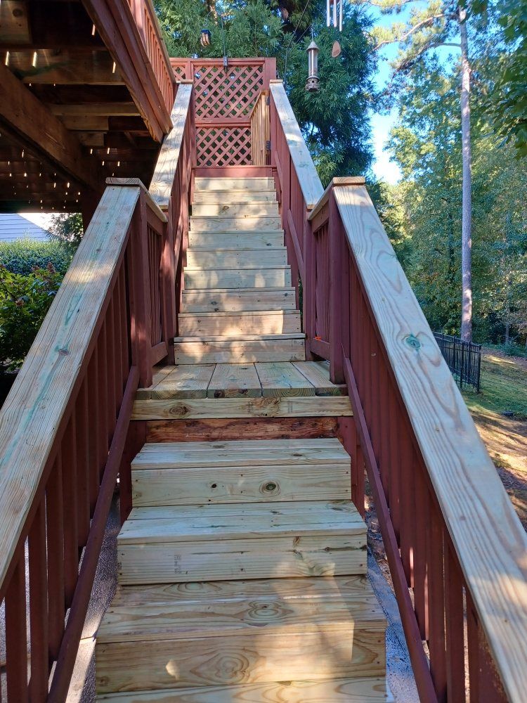 Wooden outdoor staircase with red railing leading up to a deck, set against trees and sky.