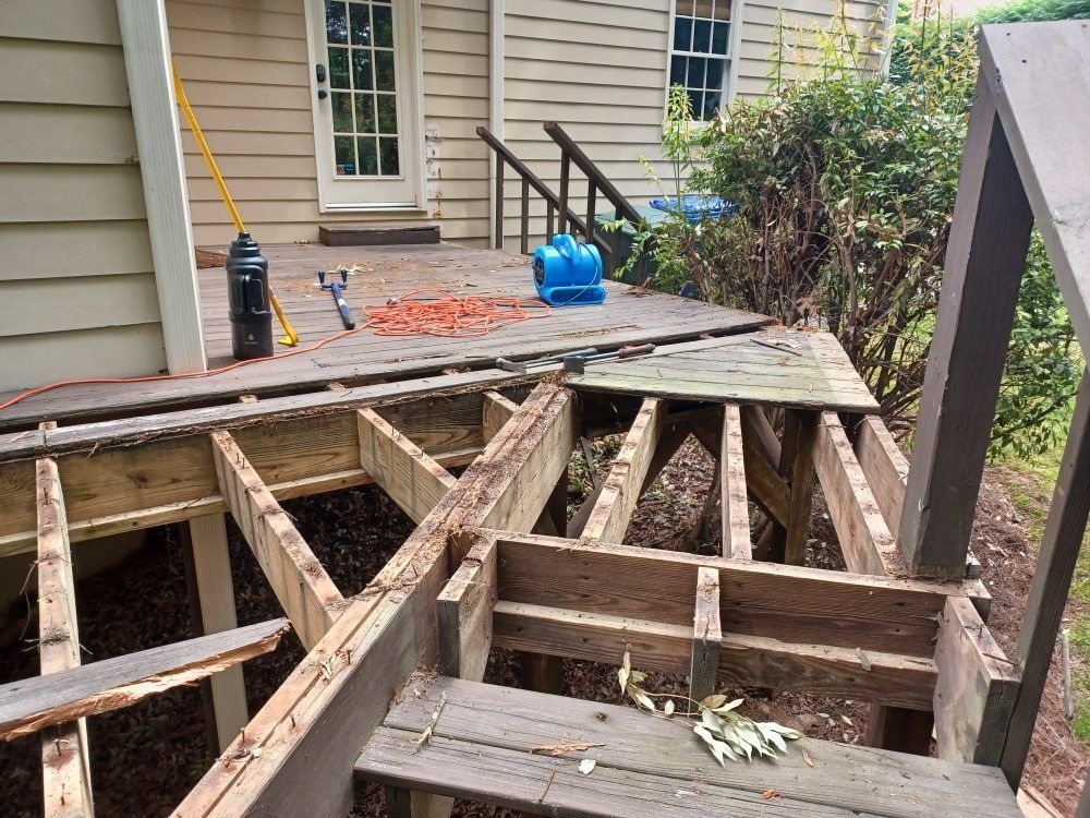 Deck under repair; exposed wooden frame with removed planks, tools, and house in background.