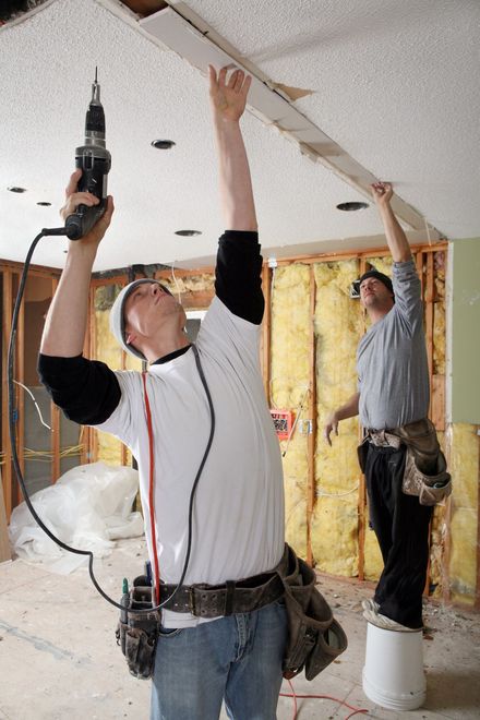 Two construction workers installing a ceiling. One drills, the other steadies a board.