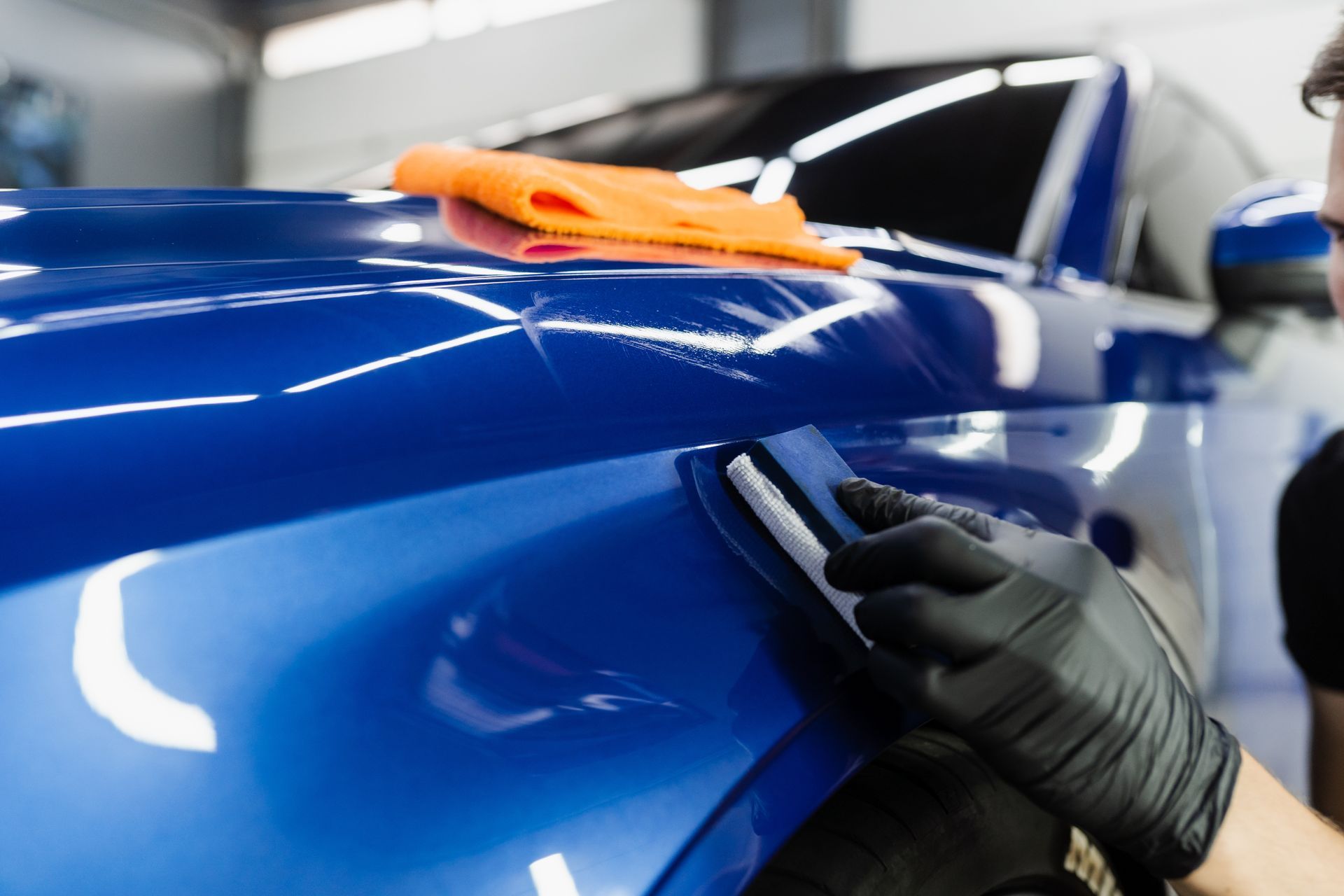 A person wearing black gloves uses a squeegee to apply a transparent protective film to a glossy blue car fender.