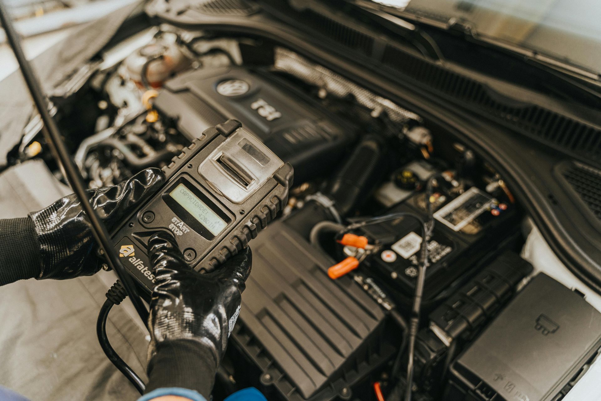 A mechanic wearing patterned gloves uses a handheld electronic diagnostic tester on a car battery inside an engine bay.