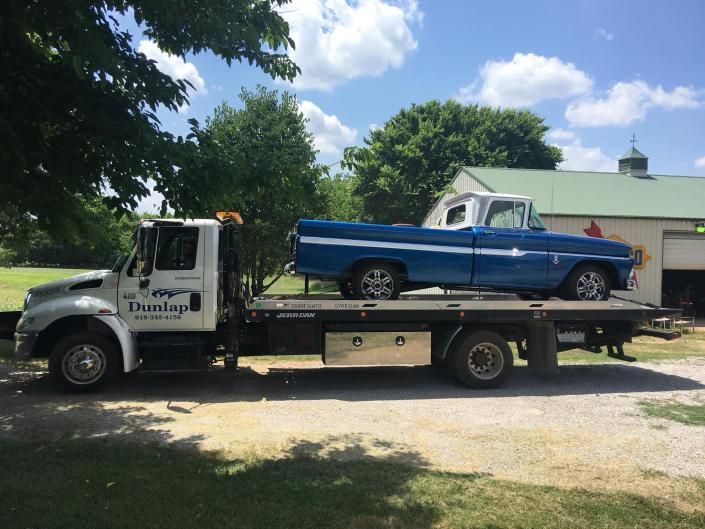 Blue and white vintage pickup truck on a flatbed tow truck in front of a barn on a sunny day.