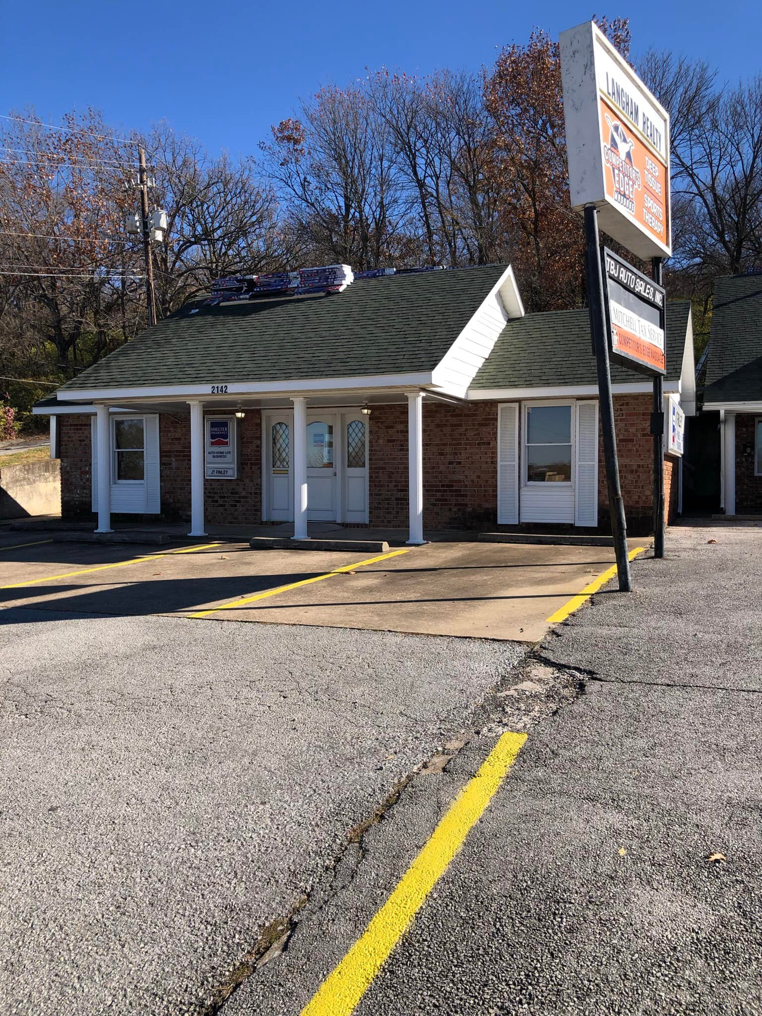 Small roadside storefront with white columns, green roof, and a sign by an empty gravel parking lot.
