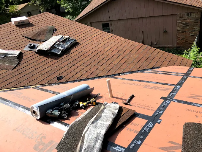 Roof under repair with orange shingles and roofing materials on top, near a house and garage