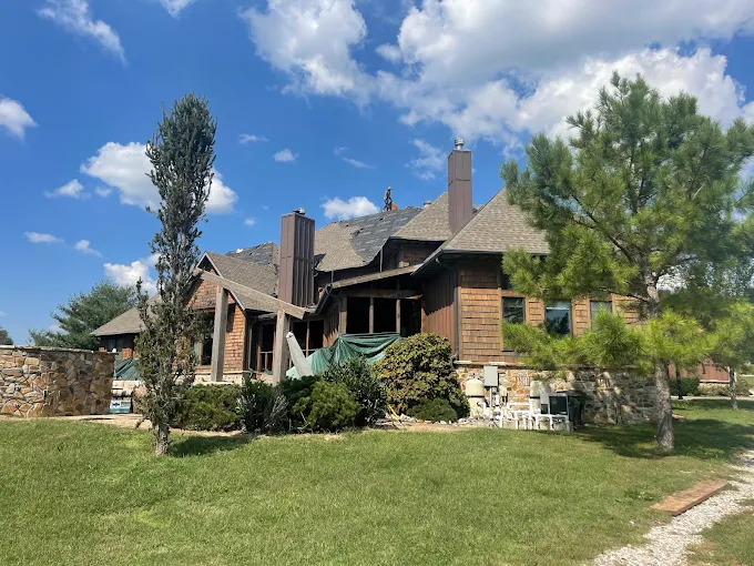 Large rustic house with brick and stone exterior, green lawn, trees, and blue sky with clouds.